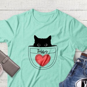 May include: A light green t-shirt with a black cat peeking out of a pocket. The pocket has a red heart with the word "Toby" above it.