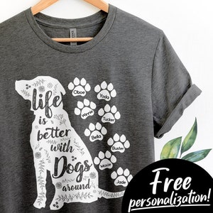 May include: A gray t-shirt with a white silhouette of a dog and the text "life is better with dogs around".  There are paw prints with dog names written inside each paw print: Oboe, Yoda, Winnie, Bella, Tucker, Buster, Moxie, and Jade.  The text "Free personalization! Add Your Dog Names" is written in a circle on the bottom right of the shirt.
