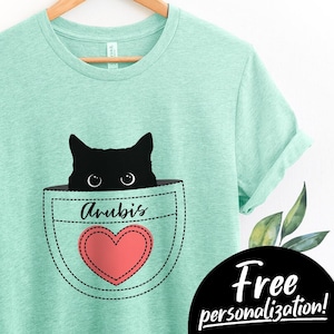 May include: A teal blue t-shirt with a black cat peeking out of a pocket with a red heart and the name "Anubis" written on it. The t-shirt is customizable with a free personalization option to add your cat's name.