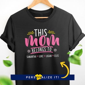 K&ouml;nnte beinhalten: Schwarzes T-Shirt mit dem Text "This Mom Belongs To" in Pink und Gelb. Unter dem Text stehen die Namen Samantha, Luke, Logan und Ellis, getrennt durch Herzen.