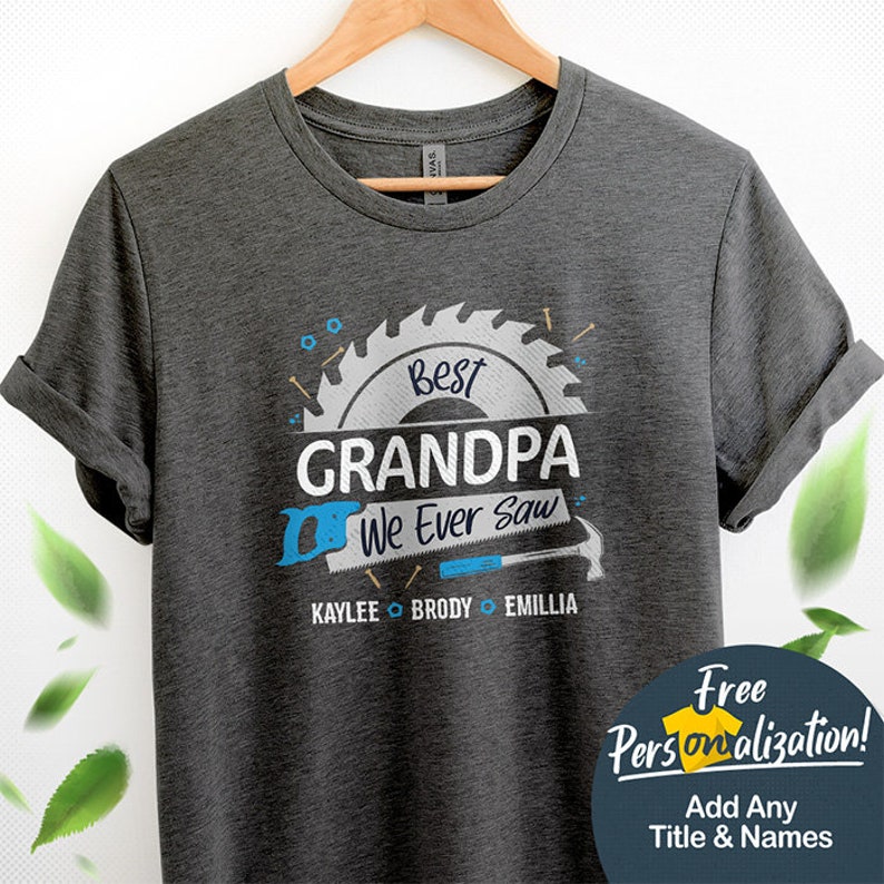 Pu&ograve; includere: Maglietta grigia con un design a forma di lama da sega blu e bianca. Il testo "Best Grandpa We Ever Saw" &egrave; all'interno della lama da sega. I nomi Kaylee, Brody ed Emillia sono sotto la lama da sega. Il testo "Free Personalization! Add Any Title & Names" &egrave; in un riquadro bianco con un'icona di camicia blu.