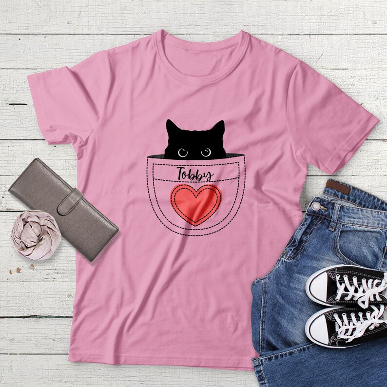 May include: Pink t-shirt with a black cat peeking out of a pocket with a red heart and the name "Toby" written on it.