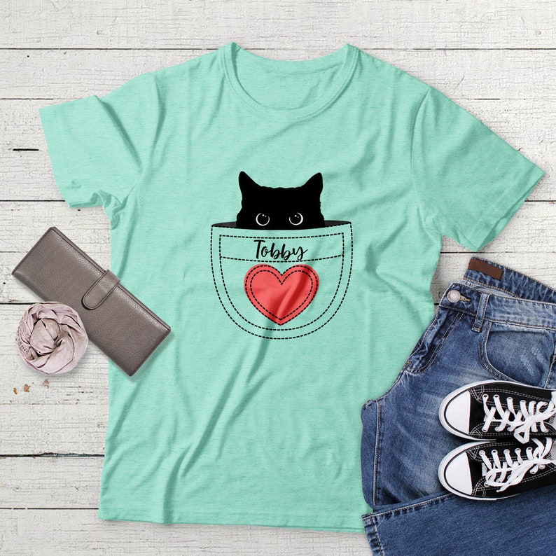 May include: A light green t-shirt with a black cat peeking out of a pocket. The pocket has a red heart with the word "Toby" above it.