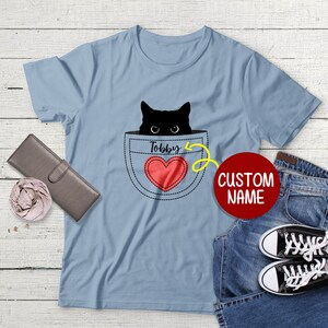 May include: A light blue t-shirt with a black cat peeking out of a pocket. The pocket has a red heart with the name "Tobby" above it. The text "CUSTOM NAME" is in a red circle.