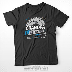 Pu&ograve; includere: Maglietta nera con una grafica di sega e martello. Il testo "Best Grandpa We Ever Saw" &egrave; scritto in bianco con una lama da sega blu dietro. I nomi "Kaylee, Brody ed Emillia" sono scritti sotto la grafica.