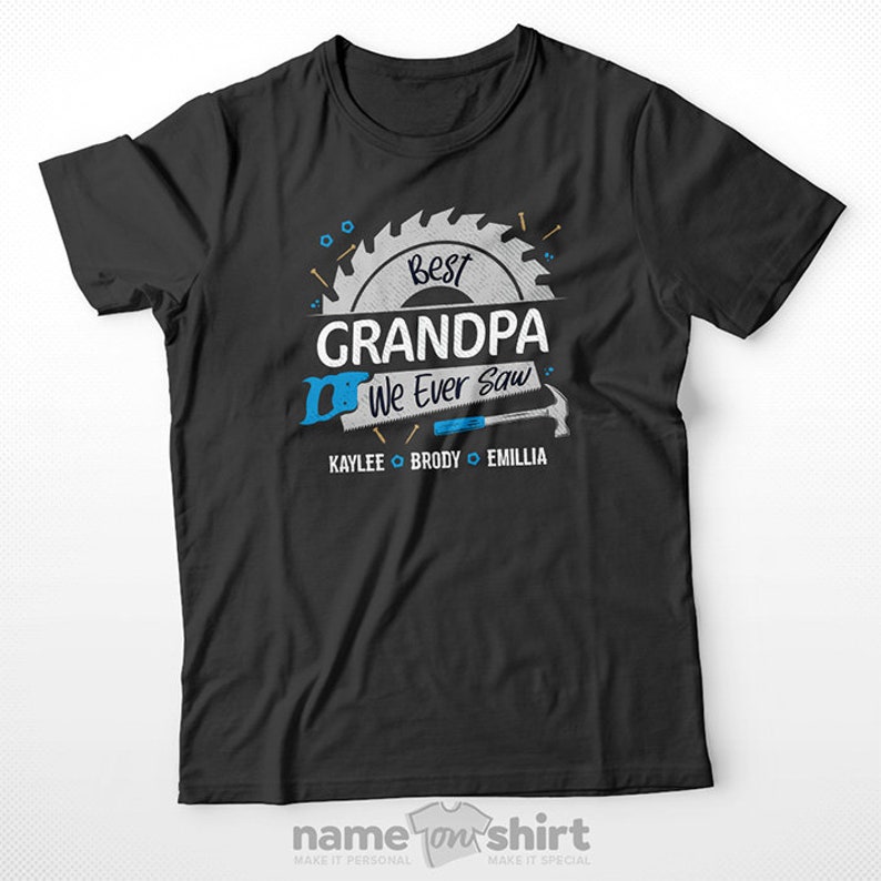 Pu&ograve; includere: Maglietta nera con una grafica di sega e martello. Il testo "Best Grandpa We Ever Saw" &egrave; scritto in bianco con una lama da sega blu dietro. I nomi "Kaylee, Brody ed Emillia" sono scritti sotto la grafica.