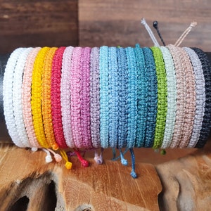 Könnte beinhalten: Eine Sammlung bunter Freundschaftsbänder aus geflochtener Schnur. Die Armbänder sind übereinander gestapelt und auf einer Holzoberfläche ausgestellt.