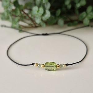 Könnte beinhalten: Ein zartes Armband aus grünem Peridot-Edelstein mit goldenen Perlen auf einer schwarzen Schnur.
