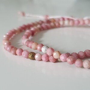 Puede incluir: Dos pulseras de piedras preciosas rosas con detalles dorados y plateados. Las pulseras est&aacute;n hechas de peque&ntilde;as cuentas facetadas y est&aacute;n atadas juntas con un cord&oacute;n blanco delgado.