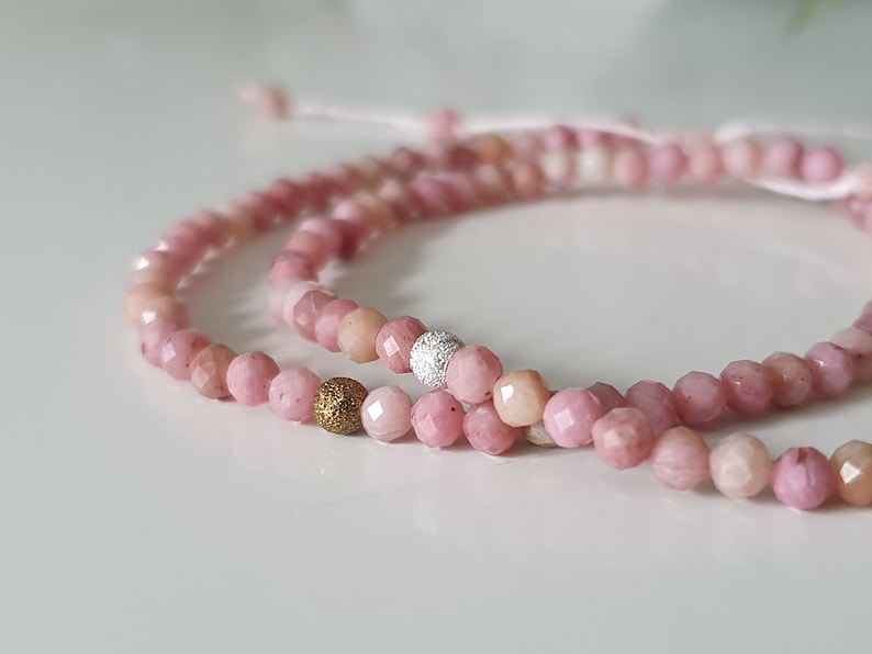 Puede incluir: Dos pulseras de piedras preciosas rosas con detalles dorados y plateados. Las pulseras est&aacute;n hechas de peque&ntilde;as cuentas facetadas y est&aacute;n atadas juntas con un cord&oacute;n blanco delgado.