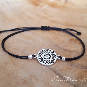 Puede incluir: Un brazalete de cuerda negra con un dije circular plateado que presenta un delicado y complejo diseño floral. El dije está acentuado con dos pequeñas cuentas plateadas.