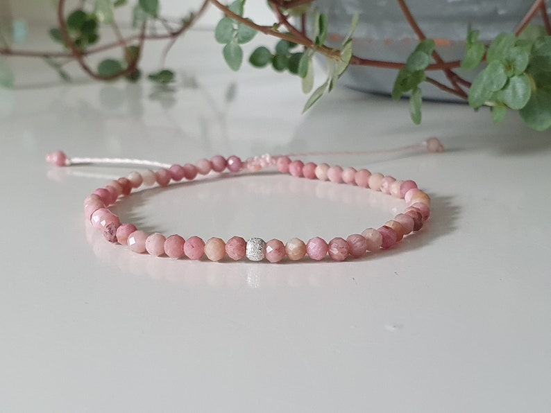 Puede incluir: Un brazalete de piedras preciosas rosas con un acento de cuentas plateadas. El brazalete est&aacute; hecho de peque&ntilde;as cuentas redondas y est&aacute; atado con un hilo delgado.