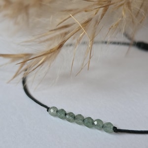 May include: A delicate bracelet featuring small, round, pale green beads strung on a thin black cord. The beads are evenly spaced, creating a minimalist design. The background includes soft, blurred, beige-colored plant stems.