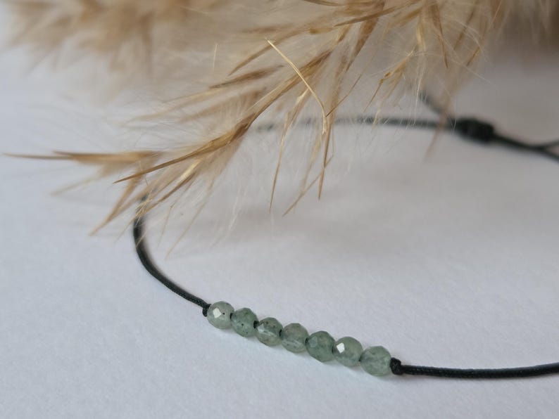 May include: A delicate bracelet featuring small, round, pale green beads strung on a thin black cord. The beads are evenly spaced, creating a minimalist design. The background includes soft, blurred, beige-colored plant stems.