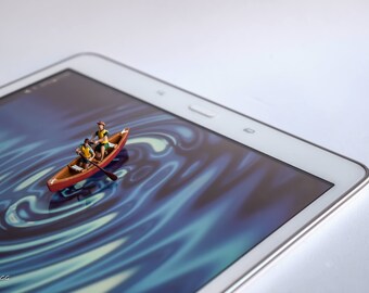 以下が含まれることがあります： 画面に青と緑の渦巻き模様が描かれた白いタブレットコンピュータ。画面上の赤いカヌーに乗ったミニチュアの人形が3人、水を漕いでいる。