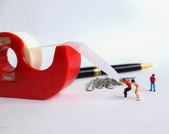 以下が含まれることがあります： 赤いテープディスペンサーと透明なテープのロール。2体のミニチュアフィギュアがテープを引っ張っており、もう1体のフィギュアが近くに立っています。画像には黒いペンと銀色のクリップも写っています。