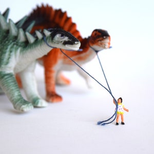 May include: A miniature person in a yellow and white dress is holding strings attached to two toy dinosaurs. The dinosaurs are green and brown with spikes and are walking forward.