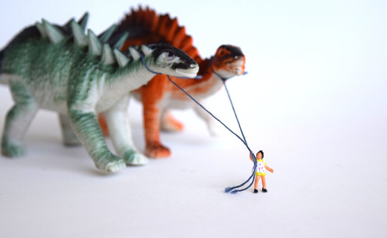 K&ouml;nnte beinhalten: Eine Miniaturperson in einem gelben und wei&szlig;en Kleid h&auml;lt F&auml;den, die an zwei Spielzeug-Dinosauriern befestigt sind. Die Dinosaurier sind gr&uuml;n und braun mit Stacheln und gehen vorw&auml;rts.
