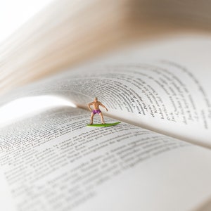 Op de afbeelding: Een miniatuurfiguur van een persoon in zwembroek die op een surfboard staat, met het surfboard op de pagina's van een open boek. Het boek is open op een pagina met tekst, en de pagina's zijn omhoog gekruld om op een golf te lijken.