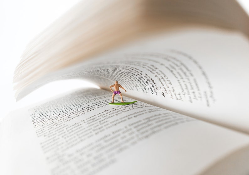 Op de afbeelding: Een miniatuurfiguur van een persoon in zwembroek die op een surfboard staat, met het surfboard op de pagina's van een open boek. Het boek is open op een pagina met tekst, en de pagina's zijn omhoog gekruld om op een golf te lijken.