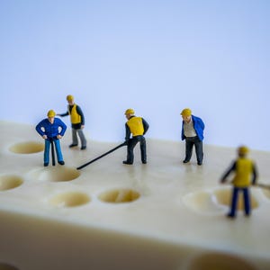 Peut inclure: Un groupe de travailleurs de la construction miniatures portant des casques jaunes et des vêtements de travail bleus travaillent sur un gros bloc de fromage suisse. Ils utilisent des outils pour creuser des trous dans le fromage.
