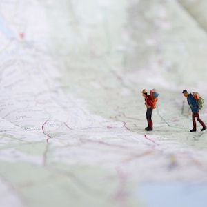 Puede incluir: Dos figuras en miniatura de excursionistas con mochilas caminando sobre un mapa detallado. Uno lleva una chaqueta roja y el otro una azul. El mapa es de color claro con carreteras y elementos geográficos.
