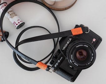 May include: A black YASHICA camera with a black leather strap and orange accents. The camera has a YASHICA LENS 50mm 1:1.8 lens. The camera is on a white surface with a roll of orange string and two rolls of film.