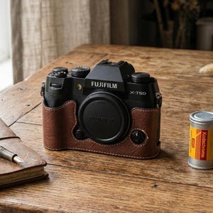 Könnte beinhalten: Eine schwarze Fujifilm X-T50 Kamera mit brauner Lederhülle liegt auf einem Holztisch neben einem Notizbuch und einem Stift. Eine Rolle Kodachrome-Film und eine Schachtel Portra 400-Film sind ebenfalls vorhanden. Auf der Kamera steht "FUJIFILM X-T50".