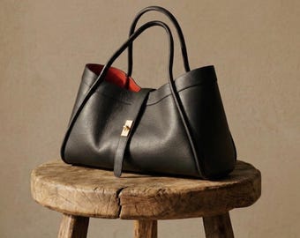 Peut inclure: Un sac fourre-tout en cuir noir avec un intérieur rouge est posé sur un tabouret en bois rustique. Le sac a deux anses et un fermoir doré. Le tabouret a trois pieds et un dessus rond, avec le grain du bois visible.