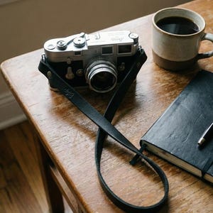 May include: A classic silver and black camera with a black leather strap sits on a wooden table. A mug of coffee, a black notebook, and a pen are also present. The image suggests a scene of traditional photography and a moment of calm.