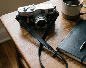 May include: A classic silver and black camera with a black leather strap sits on a wooden table. A mug of coffee, a black notebook, and a pen are also present. The image suggests a scene of traditional photography and a moment of calm.