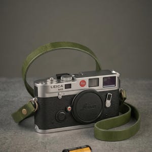 May include: A silver and black Leica M6 camera with a green leather strap and lens cap. A roll of Kodak Portra 400 film is next to it. The camera features the Leica logo and red dot.