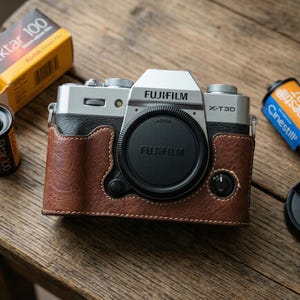 May include: A silver Fujifilm X-T30 camera with a brown leather case and lens cap. Two rolls of film, one labeled "Ektar 100" and the other "Cinestill 800T", are also present. The camera and accessories are on a wooden surface.