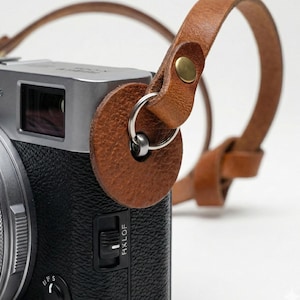 May include: A close-up of a silver and black camera with a brown leather strap attached. The strap is connected to the camera with a metal ring and a circular leather piece. The strap is secured with a brass rivet.