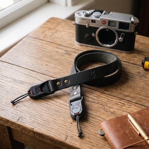 Może przedstawiać: Czarny skórzany pasek do aparatu z mosiężnymi nitami i czerwonymi akcentami leży na drewnianym stole. Srebrny i czarny aparat Leica M6, rolki filmu i skórzany notatnik z długopisem sugerują fotografię.