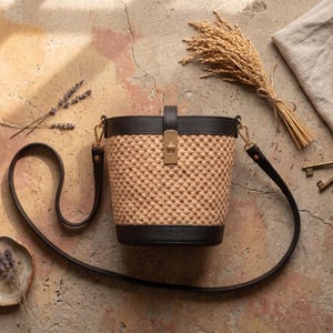 May include: A bucket bag featuring a woven tan straw body with dark brown leather trim at the top and bottom. The bag includes a gold-toned metal turn-lock closure on the front and a long, adjustable dark brown leather shoulder strap.