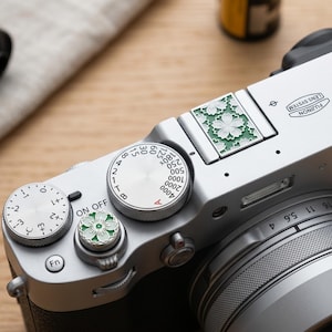 May include: A silver Fujifilm X100VI camera with a lens and dials. The camera has a green floral design on the top and a matching dial. Film rolls are visible in the background. The camera's lens has a measurement scale.