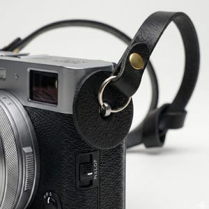Può includere: Primo piano di una tracolla per fotocamera in pelle nera attaccata a un corpo macchina argentato. La tracolla presenta un pezzo di pelle circolare con un anello di metallo e un rivetto color oro. La fotocamera ha un'impugnatura nera strutturata.