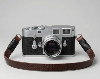 May include: A silver and black Leica camera with a brown leather strap. The camera lens is labelled "LEICA" and "LUMAR". The strap has black wrapped sections near the camera body.