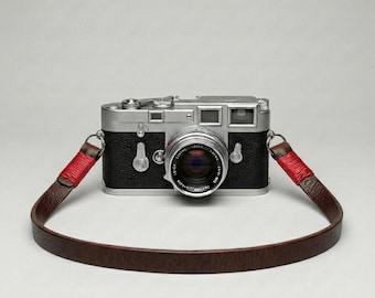 May include: A classic silver and black camera with a lens and a brown leather strap with red detailing. The camera has a vintage aesthetic, likely a film camera. The strap is attached to the camera body.