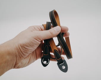 May include: A black and brown leather camera strap with orange stitching. The strap is folded in half and held in a hand.