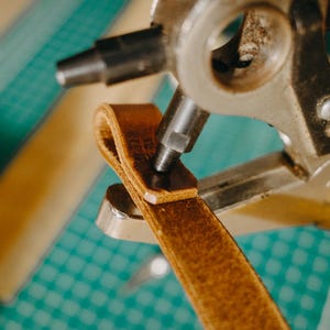 May include: A close-up of a leather strap being punched with a metal tool. The tool has a lever and a circular punch. The strap is brown and the tool is silver and black.