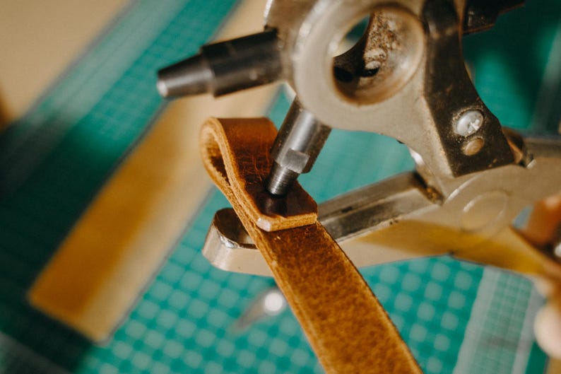 May include: A close-up of a leather strap being punched with a metal tool. The tool has a lever and a circular punch. The strap is brown and the tool is silver and black.