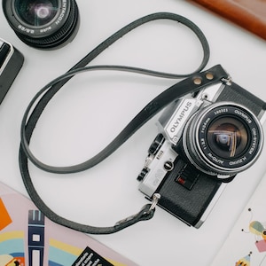May include: A silver and black Olympus camera with a black leather strap. The camera has a silver lens with a black rim and the words "OLYMPUS" and "OM-SYSTEM" printed on it.