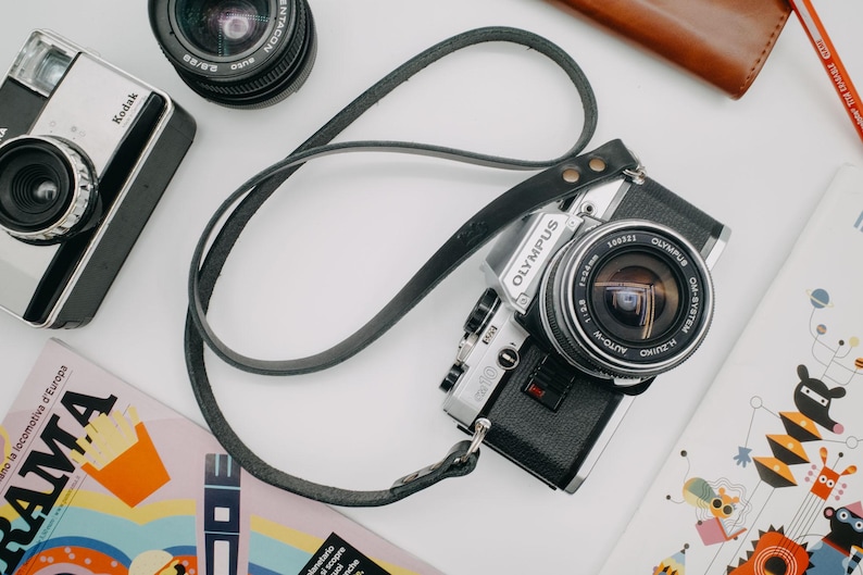 May include: A silver and black Olympus camera with a black leather strap. The camera has a silver lens with a black rim and the words "OLYMPUS" and "OM-SYSTEM" printed on it.