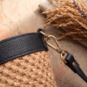May include: A woven tan straw bucket bag featuring a black leather trim along the top edge. The bag includes a detachable black leather strap connected by a brass-toned metal lobster clasp hardware, showcasing a textured finish on the leather.