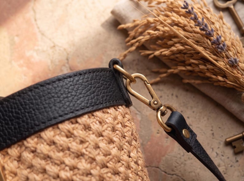 May include: A woven tan straw bucket bag featuring a black leather trim along the top edge. The bag includes a detachable black leather strap connected by a brass-toned metal lobster clasp hardware, showcasing a textured finish on the leather.