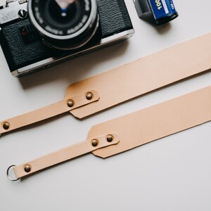 May include: A light brown leather camera strap with two metal rings and gold-coloured rivets. The strap is laid out flat on a white surface.