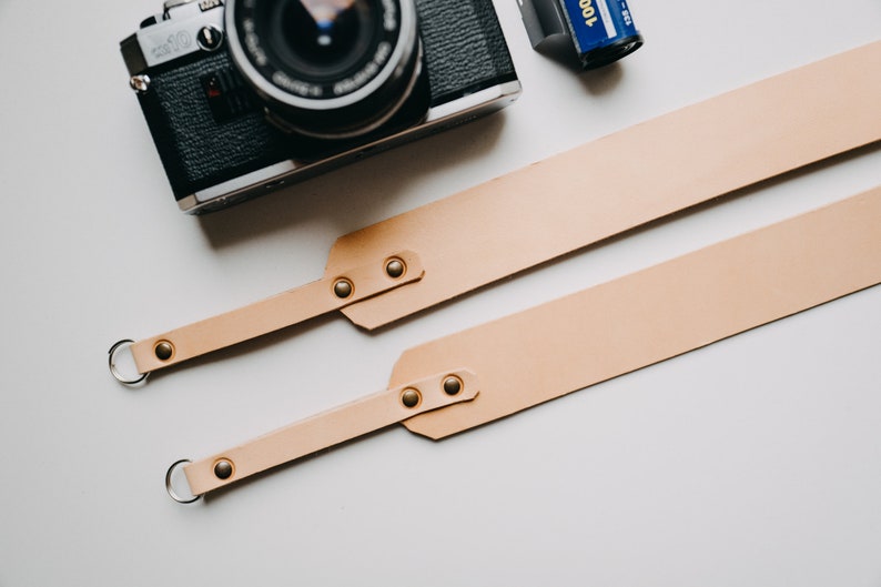May include: A light brown leather camera strap with two metal rings and gold-coloured rivets. The strap is laid out flat on a white surface.