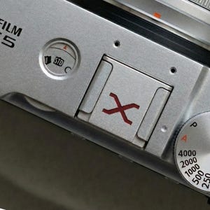 May include: Close-up of a silver Fujifilm X-E5 camera body. Features include a red 'X' mark, a dial with settings from 125 to 4000, and a dial with STD and C settings. The camera's top is visible.